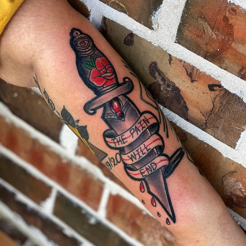 Tattoo of a dagger with roses and text on a person's arm, against a brick wall background.
