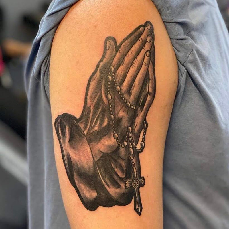 Praying hands tattoo with a rosary on a client’s bicep. 